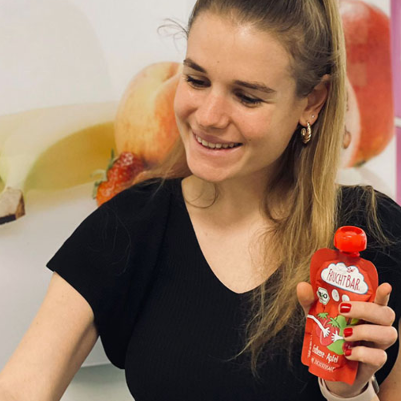 FruchtBar Mitarbeiterin lächelt und hält einen roten Quetschie hoch.