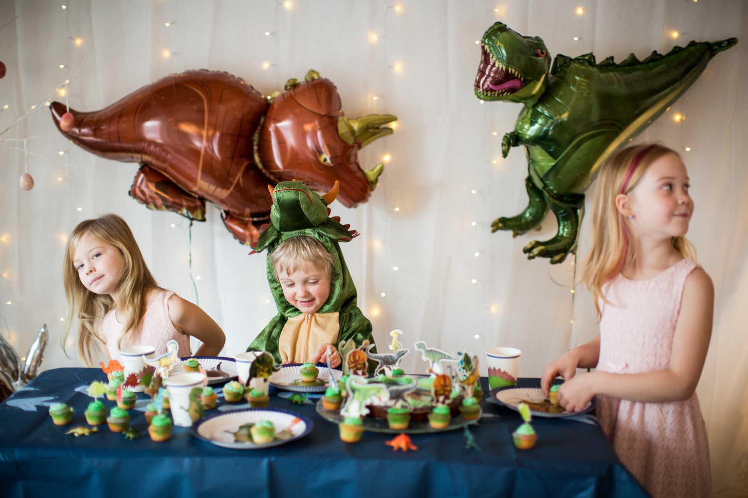 Foto von Kindern auf einer Dino-Geburtstagsfeier mit Dino-Ballons im Hintergrund und grünen Muffins auf dem Tisch 