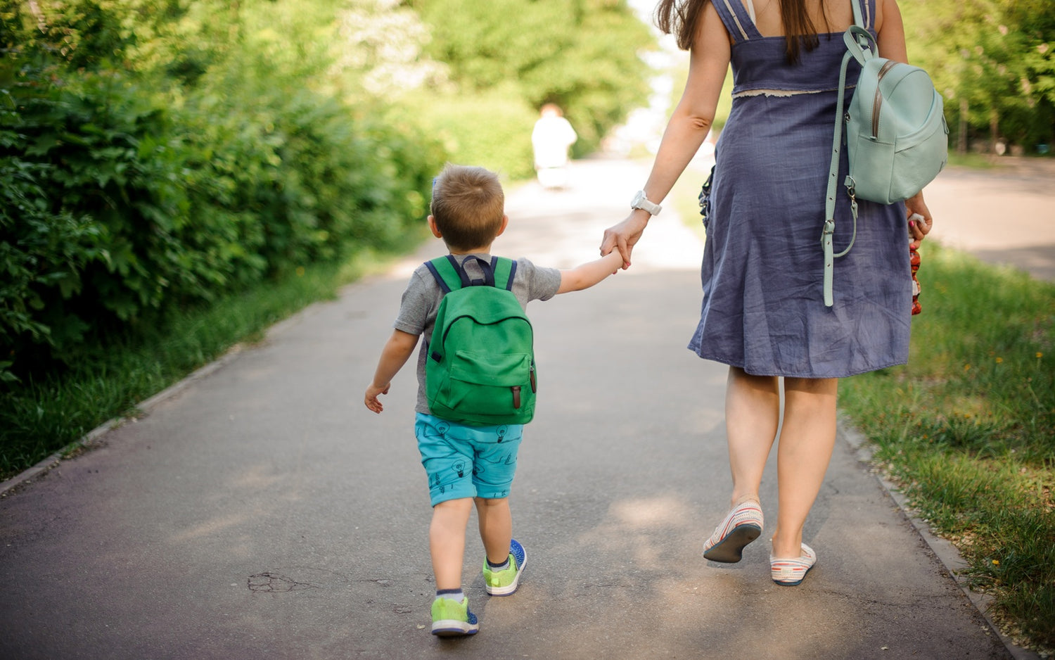 Kleiner Junge mit grünem Rucksack läuft Hand in Hand mit seiner Mama auf dem Bürgersteig