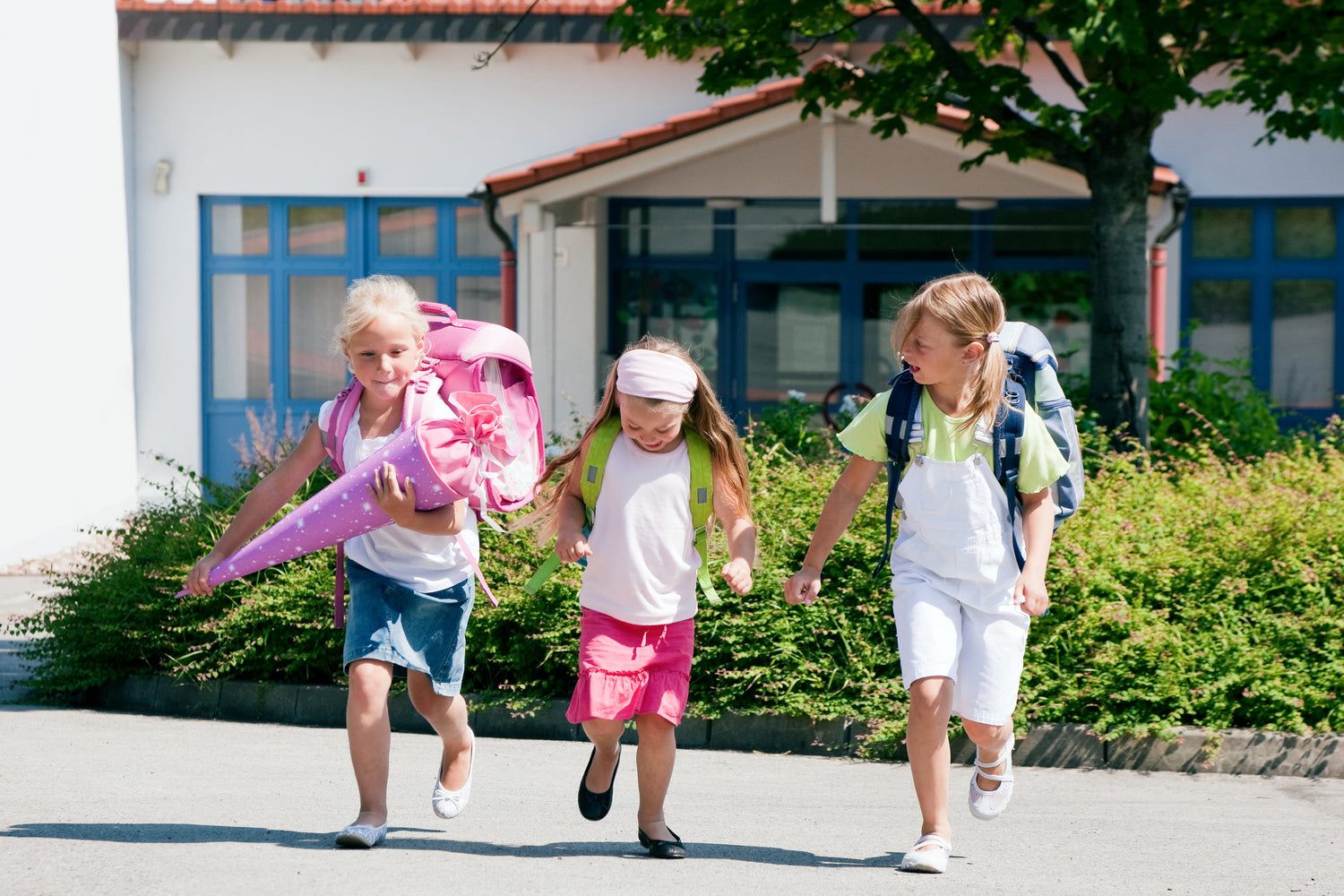 Drei Mädchen mit Schulrucksäcke laufen fröhlich auf die Kamera zu, eins hält ihre Schultüte in der Hand