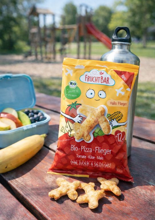 Eine Packung FruchtBar Bio Pizza-Flieger steht auf einer Bank auf einem Spielplatz neben einer Brotdose und einer Trinkflasche.