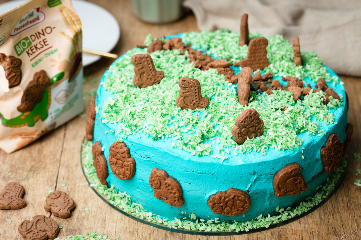 Foto einer Dino-Welt-Torte: Schokokuchen mit blauer Creme, grünen Kokosflocken als Gras und Bio Dino Keksen zur Deko.